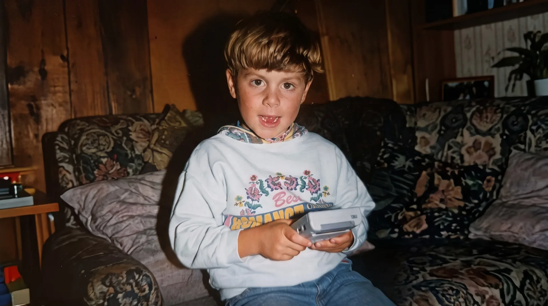 Luka als Kind sitzt mit einem Gameboy auf einem geblümten Sofa und schaut in die Kamera