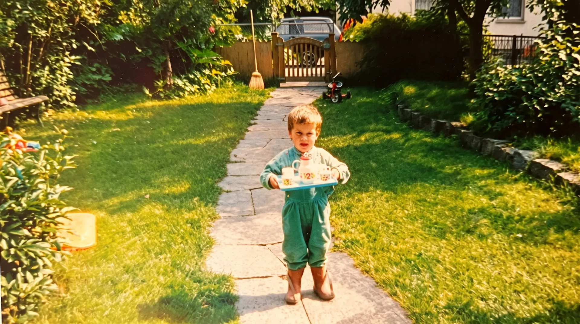 Kleiner Luka in grünem Anzug trägt ein Tablett mit Spielzeug-Teeservice über einen Gartenweg